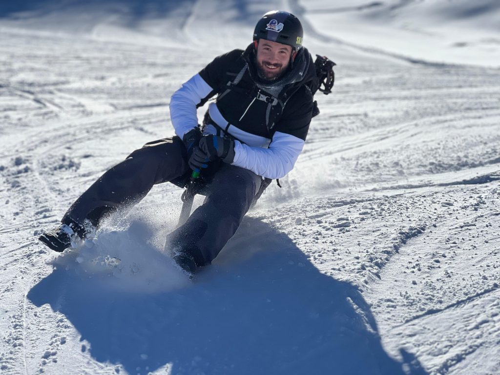 Un skieur sourit en descendant une pente enneigée avec un Snooc, portant un casque et des vêtements sombres, entouré de traces de glisse.