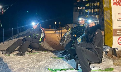 Snooc After Ski à Courchevel : Glisse Nocturne en Groupe ! Trois personnes assises sur un Snooc avec des frontales, prêtes à descendre une pente enneigée à Courchevel le soir, dans un décor alpin éclairé.